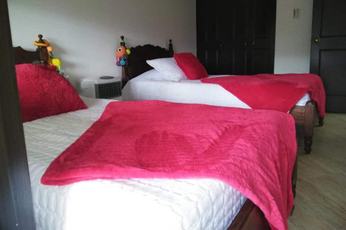 A room with two beds, each with a red blanket and white bedding. There is a small table with a lamp and a toy on it.