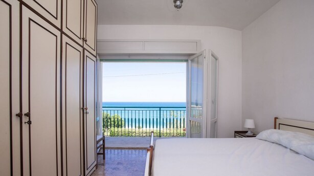 Camera da letto con un letto, un comodino, una lampada e vista sul mare attraverso una porta aperta.