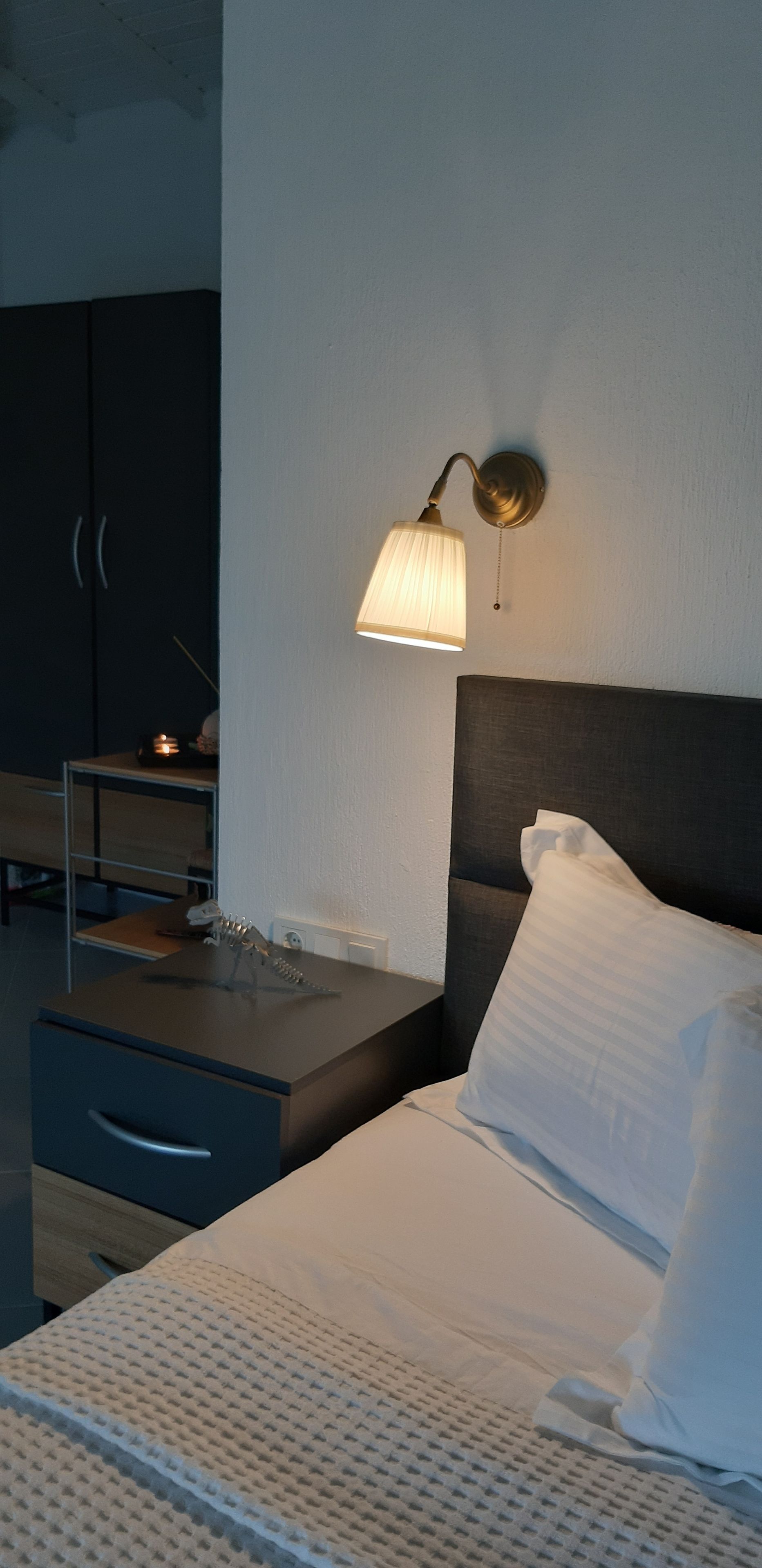 A hotel room with a bed, a bedside table, a lamp, a wardrobe, and a small desk with a lamp.