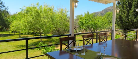 Un área de comedor al aire libre techada con una mesa preparada para dos, rodeada por una barandilla de metal, con vistas a un paisaje cubierto de césped, árboles y colinas al fondo.