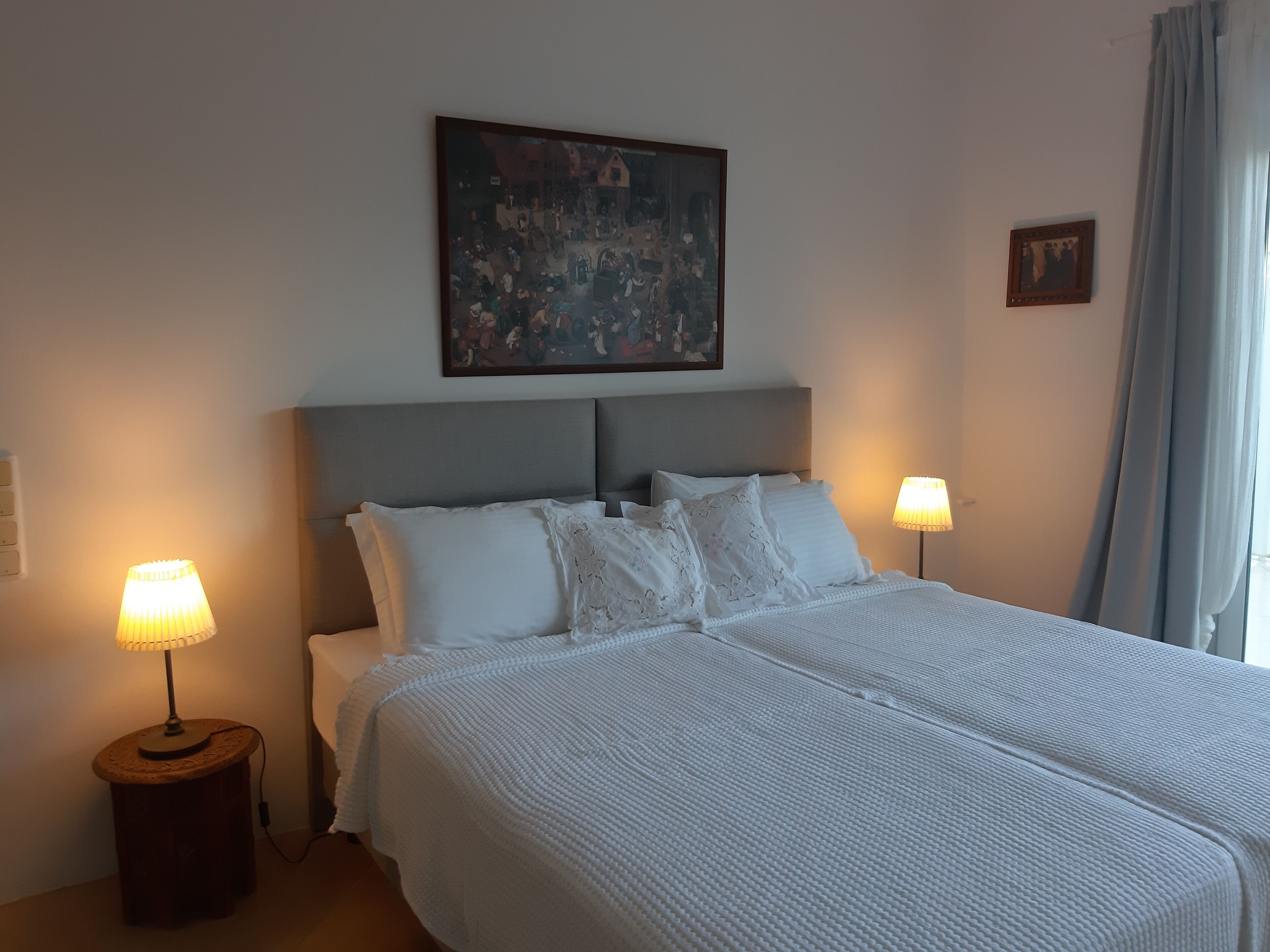 A hotel room with a large bed, two bedside lamps, a painting on the wall, and a window with curtains.