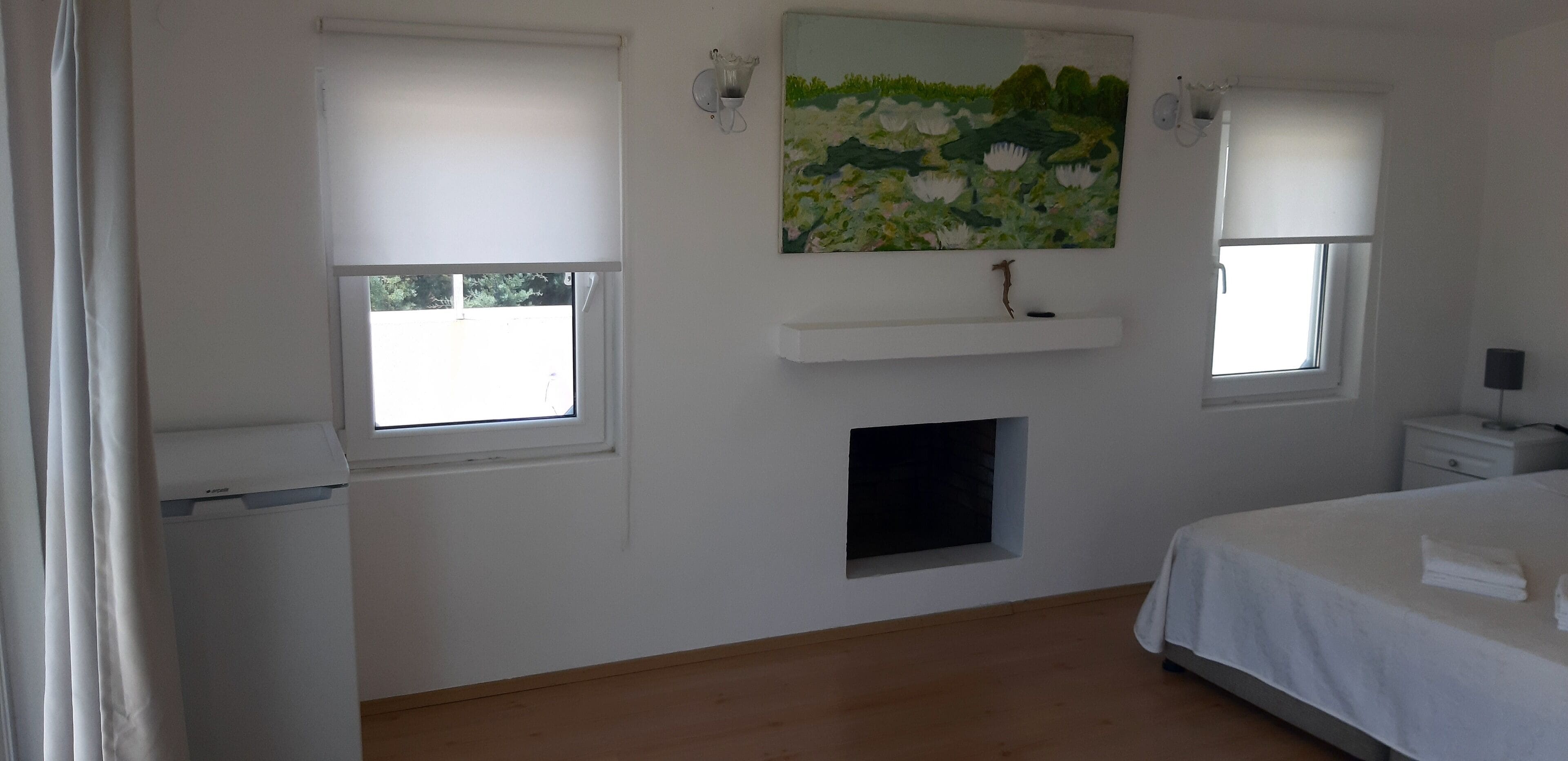 Une chambre avec deux fenêtres, un tableau, une petite étagère, une table de chevet et un lit.