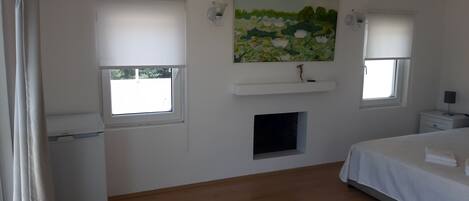 Une chambre avec deux fenêtres, un tableau, une petite étagère, une table de chevet et un lit.
