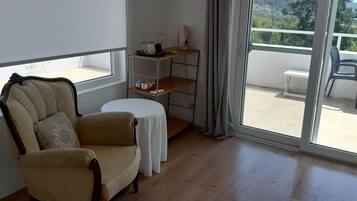 A room with a beige armchair, a small round table, a white tablecloth, a metal shelf with kitchen items, and a view of a balcony with a chair.