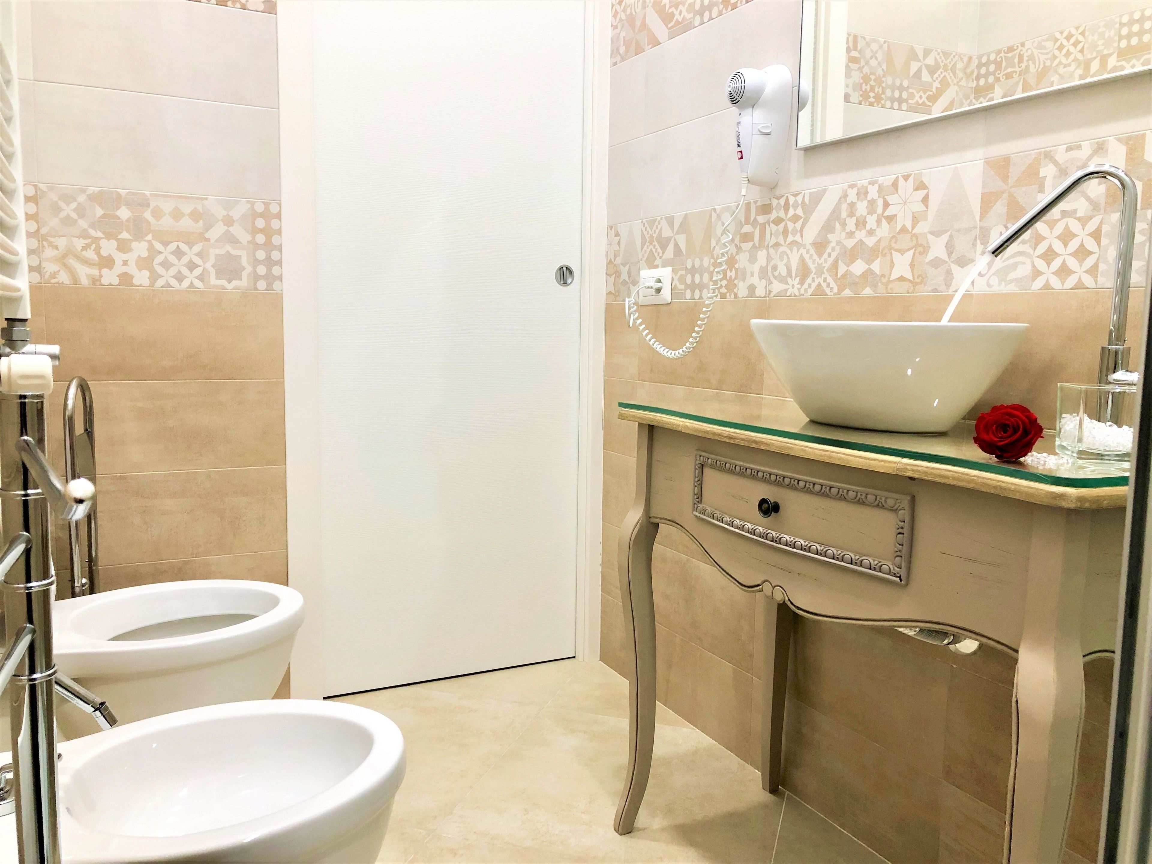 A bathroom with two toilets, a sink with a glass bowl, a hairdryer on the wall, and a decorative red rose on the counter.