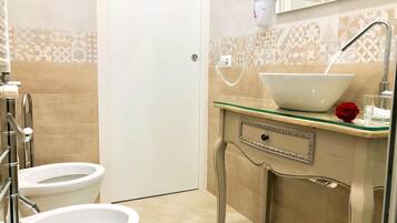 A bathroom with two toilets, a sink with a glass bowl, a hairdryer on the wall, and a decorative red rose on the counter.