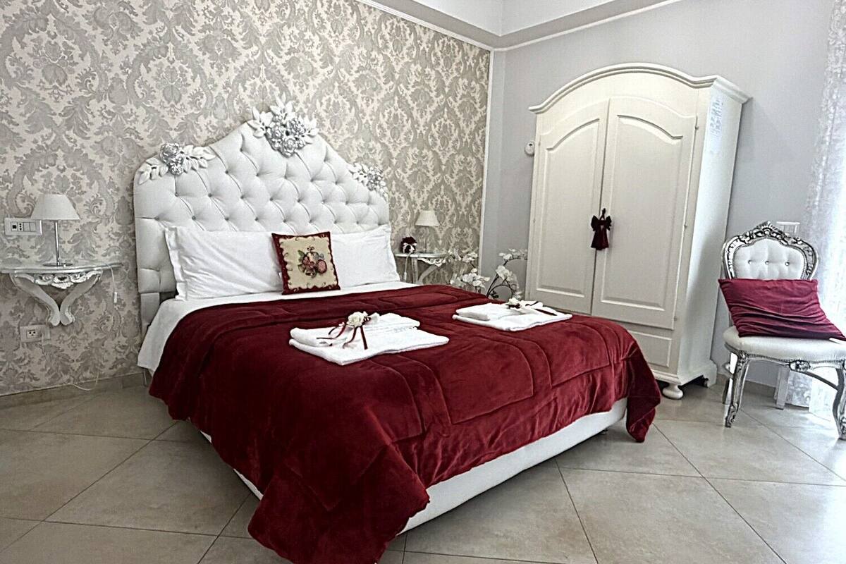 Ein Schlafzimmer mit einem großen Bett, einem weißen Kopfende und einer roten Bettdecke. Es gibt einen weißen Kleiderschrank und einen Sessel mit einem roten Kissen.