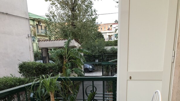 Un pequeño balcón exterior con una mesa redonda y dos sillas blancas, una planta en maceta y vistas a la calle y edificios al fondo.