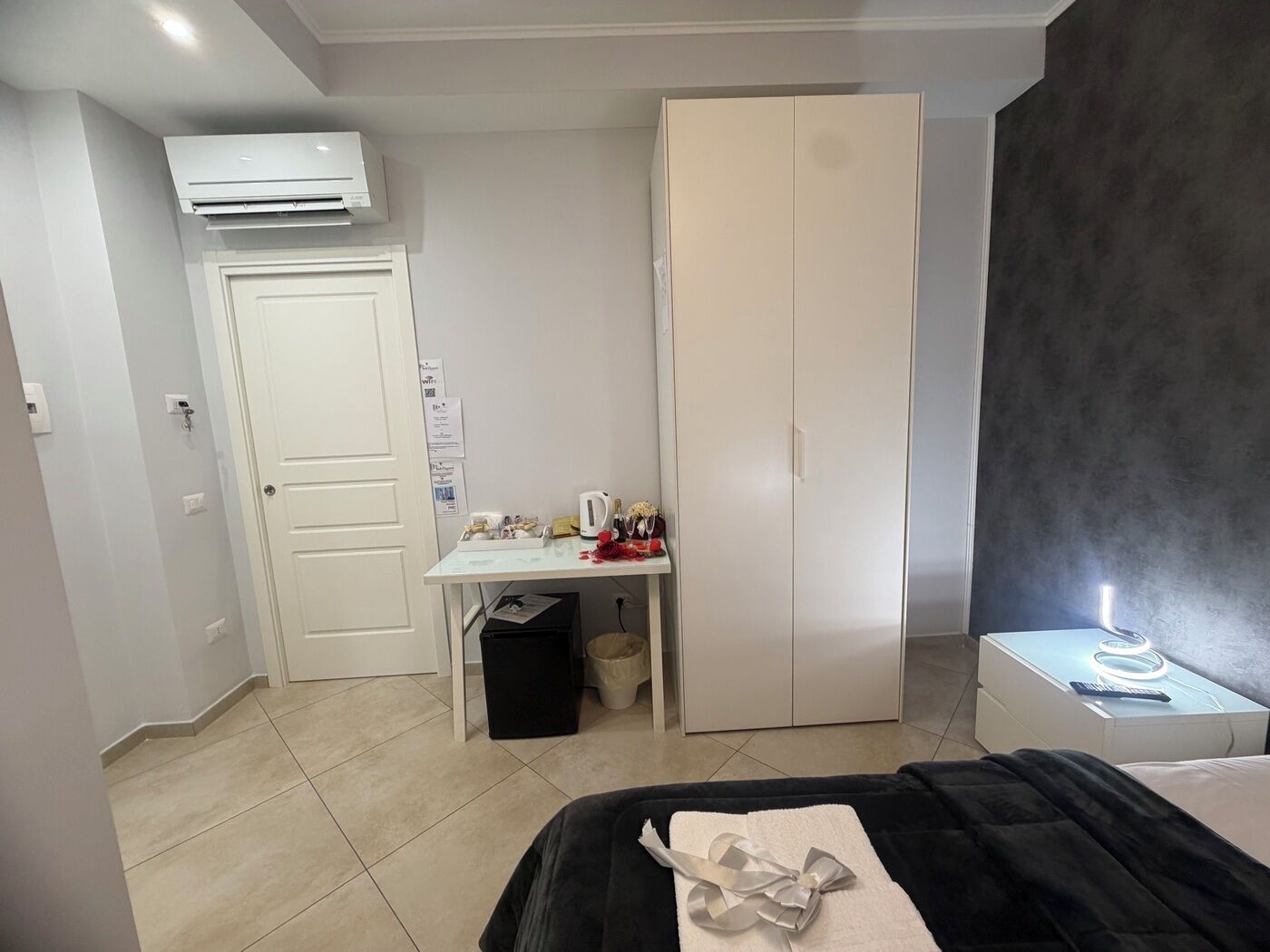 A bedroom with a bed, a desk, a wardrobe, and a bedside table with a lamp.
