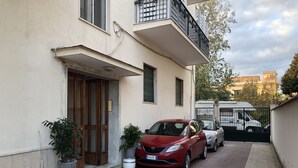 Un edificio residencial con un camino de entrada cerrado por una reja, un coche rojo estacionado al frente y una furgoneta blanca dentro de la puerta.