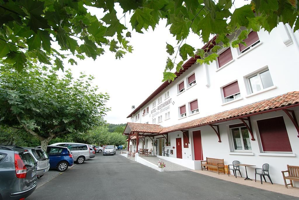 A white building with a red-tiled roof, a sign indicating it is a hotel and restaurant, and a parking area with several cars.