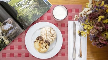 A table with a book, a plate of pastries, a glass of milk, cutlery, and a vase of flowers.