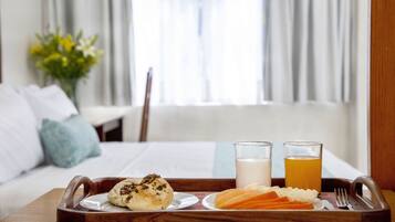 Bandeja de café da manhã com pães, frutas e dois copos de leite e suco, sobre uma bandeja de madeira em um quarto de hotel.