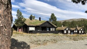 A row of black wooden cabins with grass roofs, set on a gravel ground with trees and hills in the background. - Randsverk Hytter i Lemonsjøen Fjellpark (Vaga)