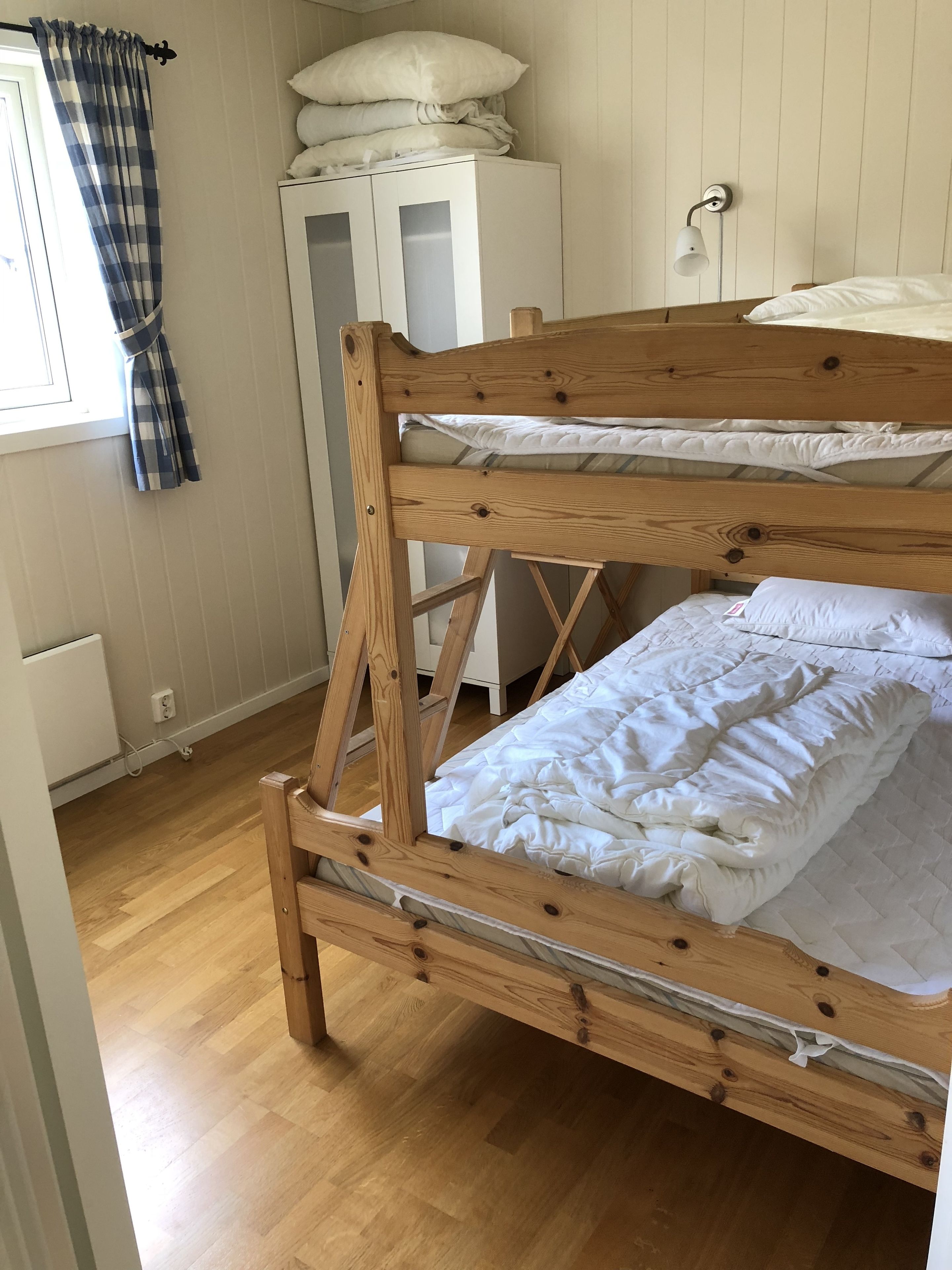 A bunk bed with wooden frames and white bedding, a white wardrobe, and a window with curtains.