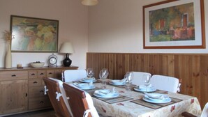 Un comedor con una mesa de madera dispuesta para cuatro personas, un armario de madera con cajones, un reloj y un cuadro en la pared.
