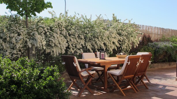 Un espacio exterior para comer con una mesa y sillas de madera, rodeado de abundante vegetación y arbustos florecientes.
