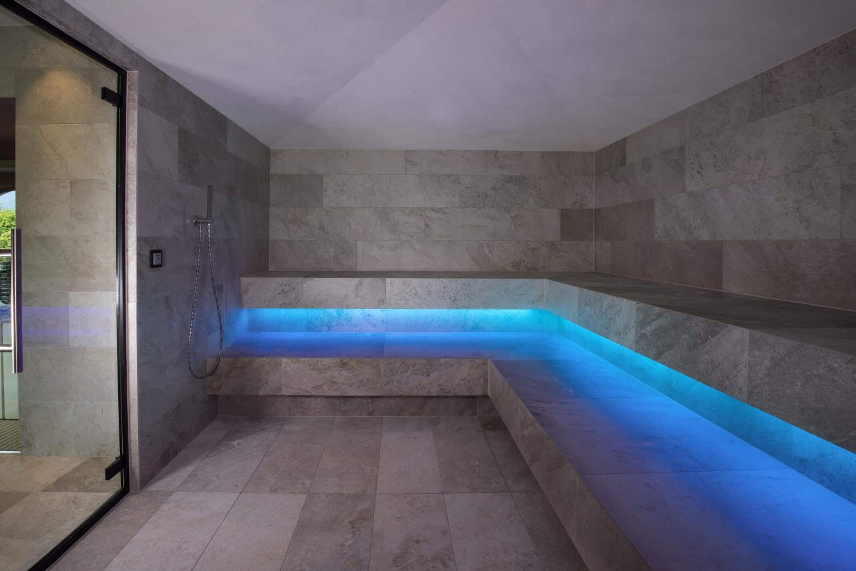 Un moderno espacio de spa con suelo calefaccionado, entrada por puerta de vidrio y fuente de agua iluminada en azul.