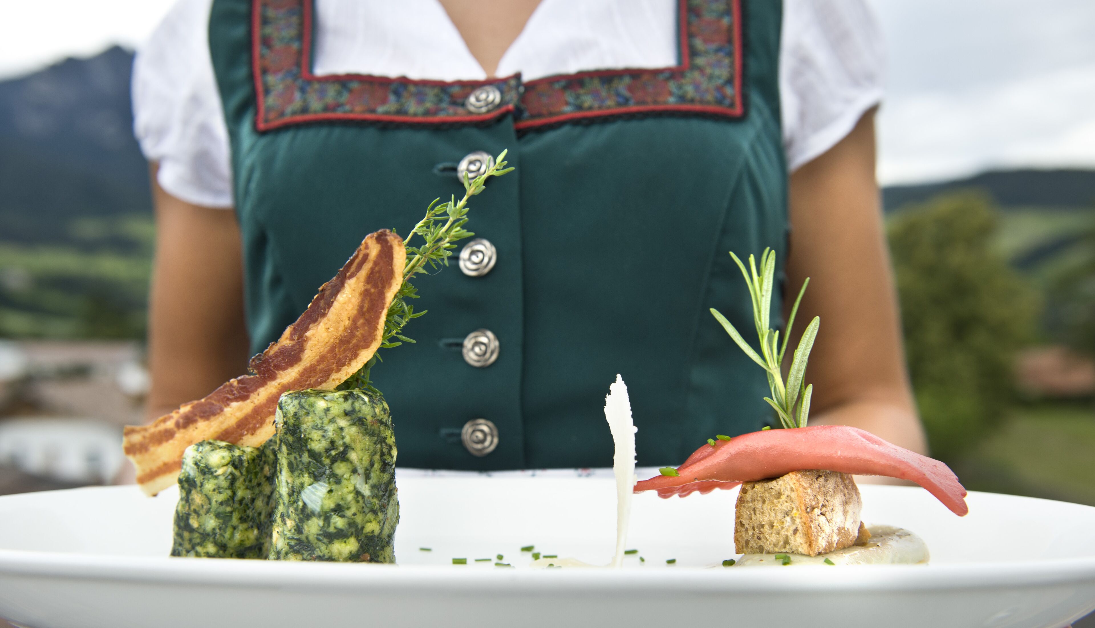 Un piatto con diversi cibi, tra cui un pezzo di carne, un formaggio verde e del bacon, con una persona in abiti tradizionali sullo sfondo.