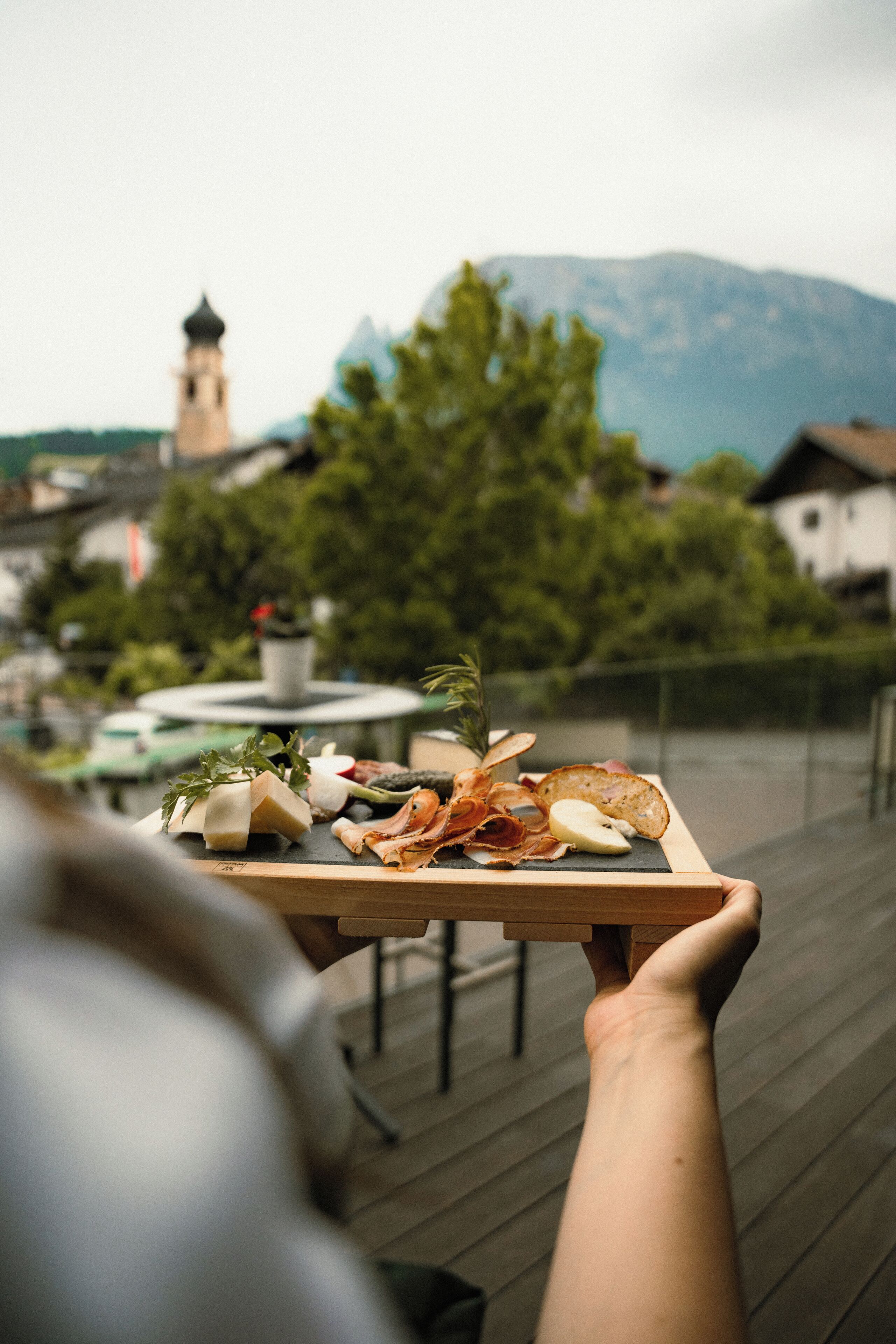 A person holding a tray with various food items, a table, and a mountain in the background.