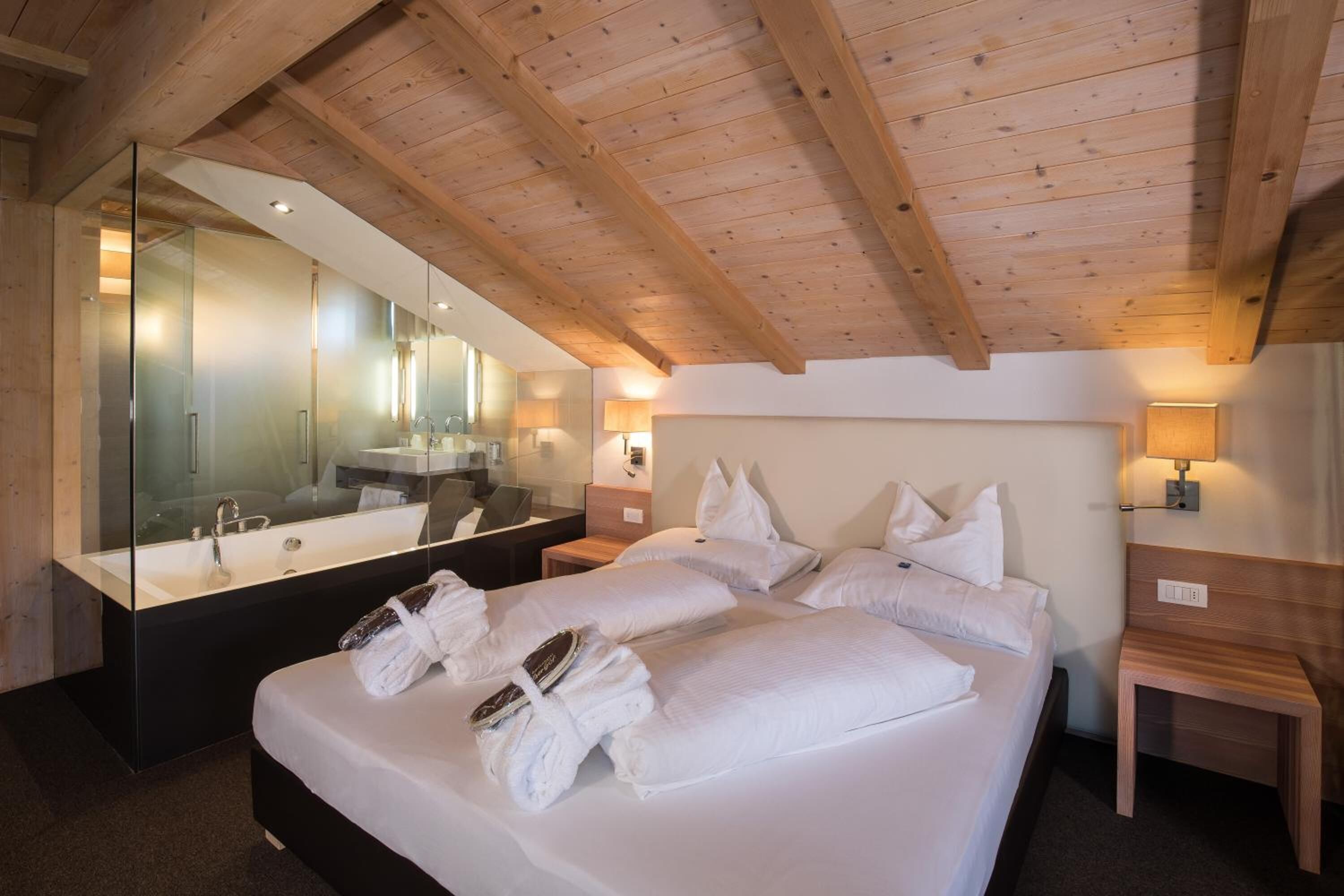 A modern hotel room with a large bed, a glass-enclosed bathroom, wooden ceiling beams, and a large mirror.