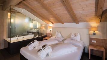 A modern hotel room with a large bed, a glass-enclosed bathroom, wooden ceiling beams, and a large mirror.