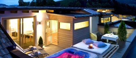 Een moderne, overdekte hottub omgeving met een houten terras, lounge stoelen en een kleine tafel met een kaars.