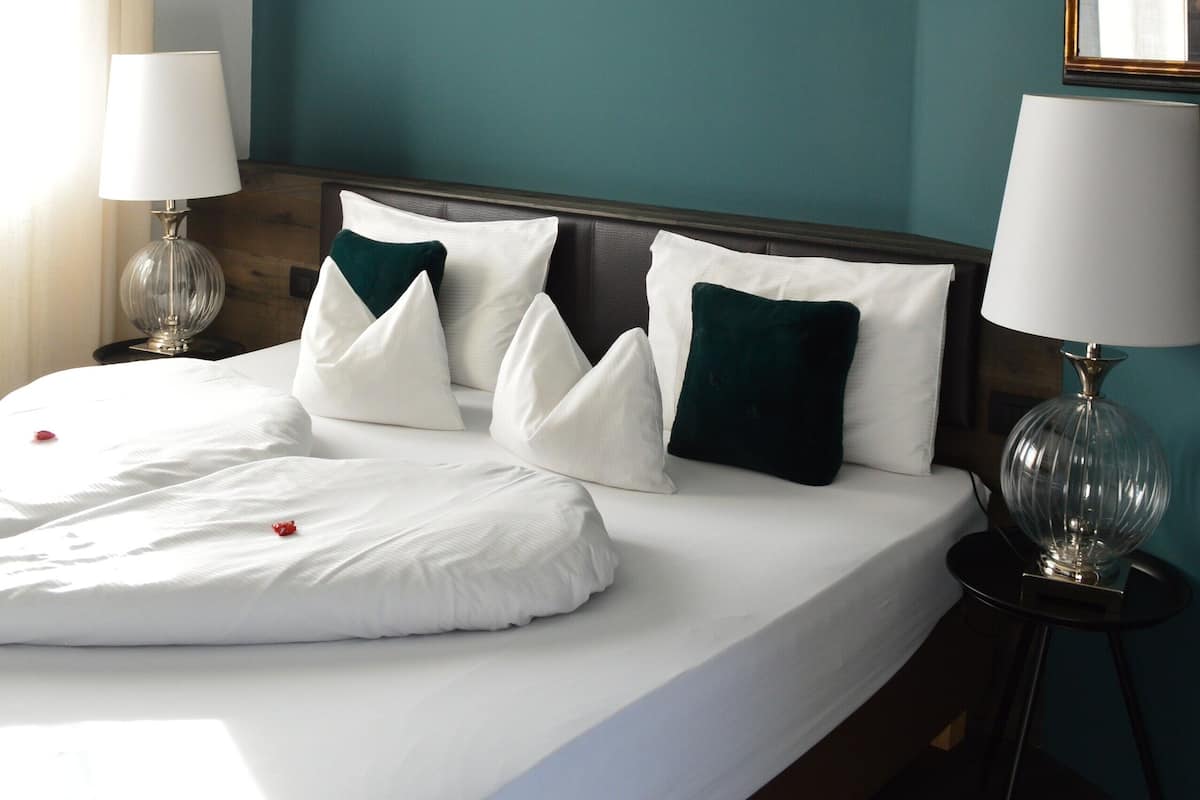 A neatly made bed with white linens and green pillows, a wooden headboard, and a chandelier above.