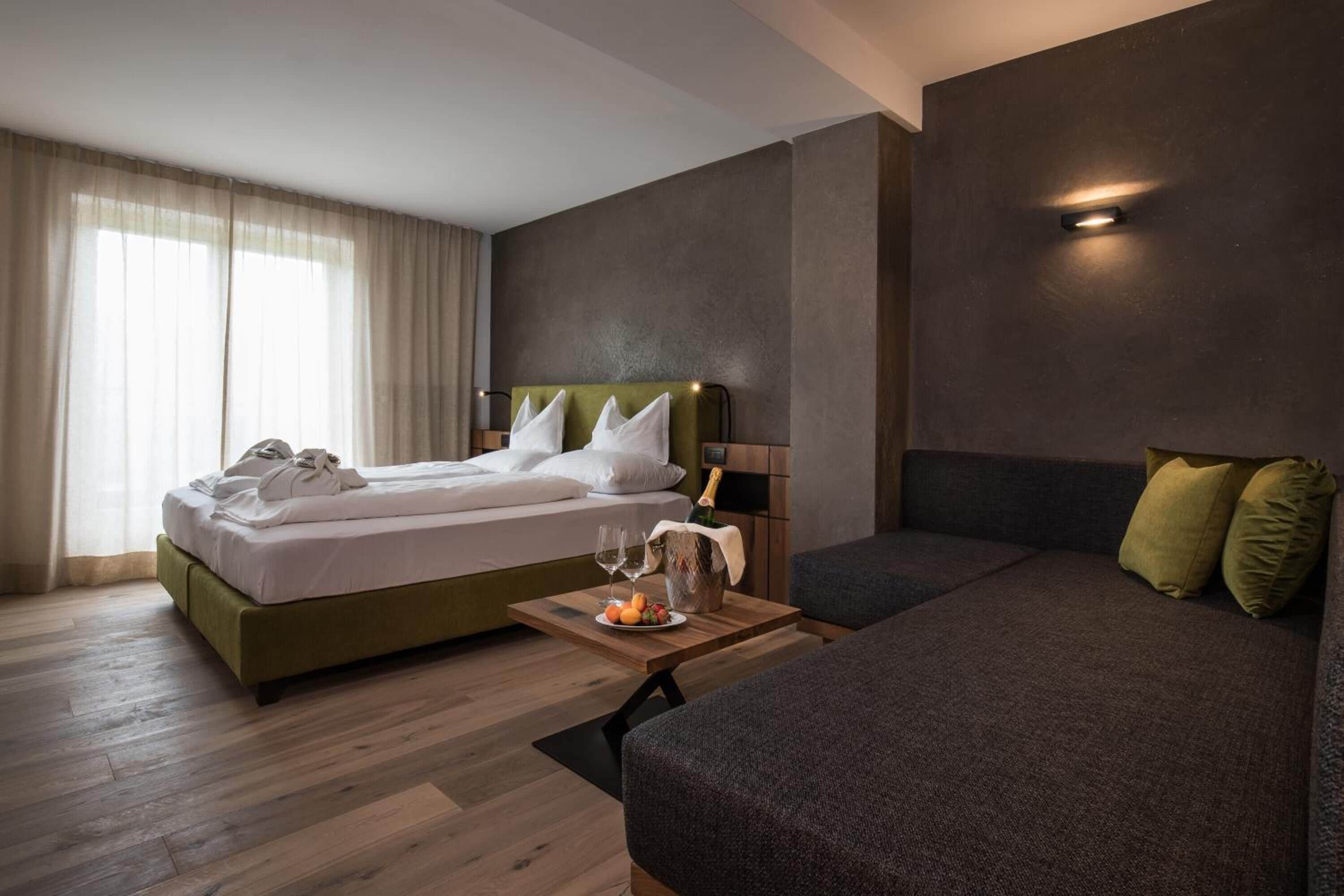 Una habitación de hotel con una cama, un sofá, una mesita con una botella y vasos, y una ventana con cortinas.