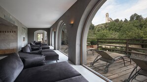 A modern interior with a long row of dark sofas and a wooden deck with chairs, overlooking a lush landscape and a distant church.