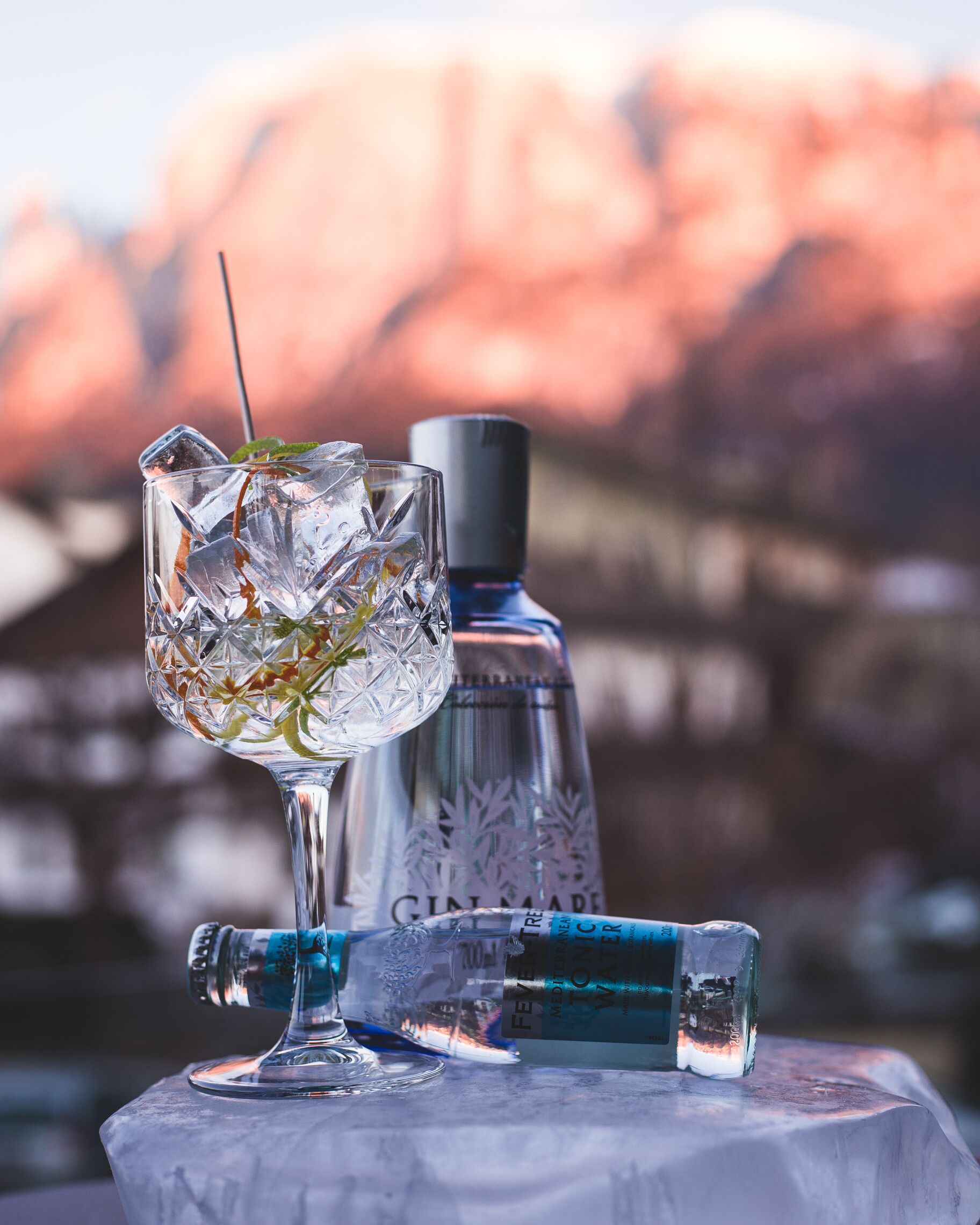 Un vaso con hielo y una hoja de menta, una botella con la etiqueta 'Gin Made', y una tapa de botella sobre una mesa con una montaña de fondo.