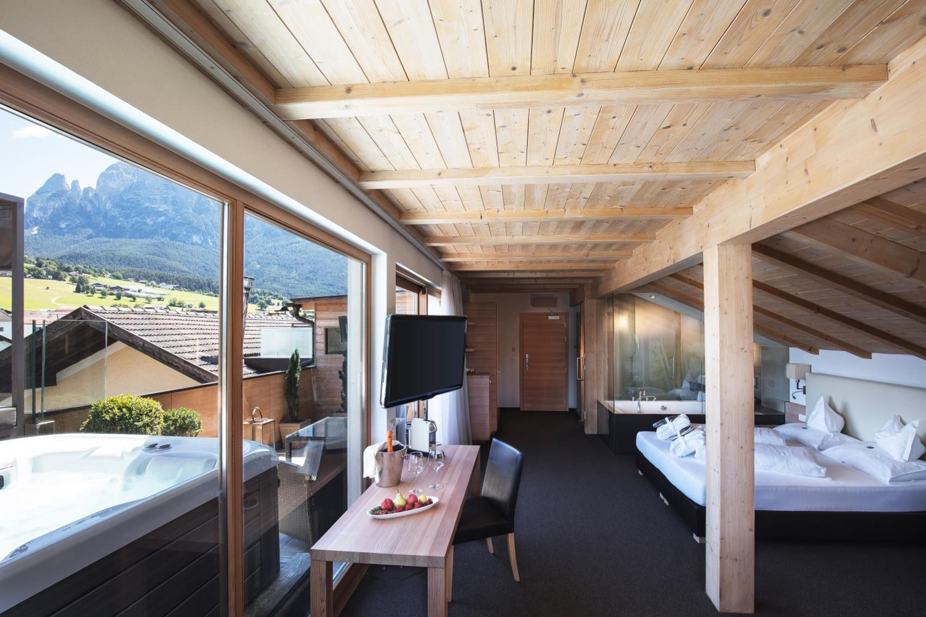 Una habitación de hotel moderna con techo de madera, un ventanal con vistas a la montaña, baño privado y zona de comedor con jacuzzi.