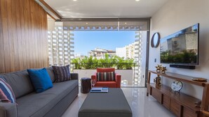 A modern living room with a sofa, ottoman, and a red chair. A TV is mounted on the wall, and there's a balcony with a view of buildings. - Legendary Mexican Architecture in the Heart of Zona Romantica! 1BD Studio for re (Puerto Vallarta)