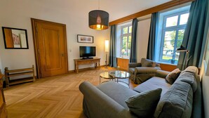 Una sala de estar con un sofá, un televisor, un banco de madera, una mesa y un ventanal con cortinas.