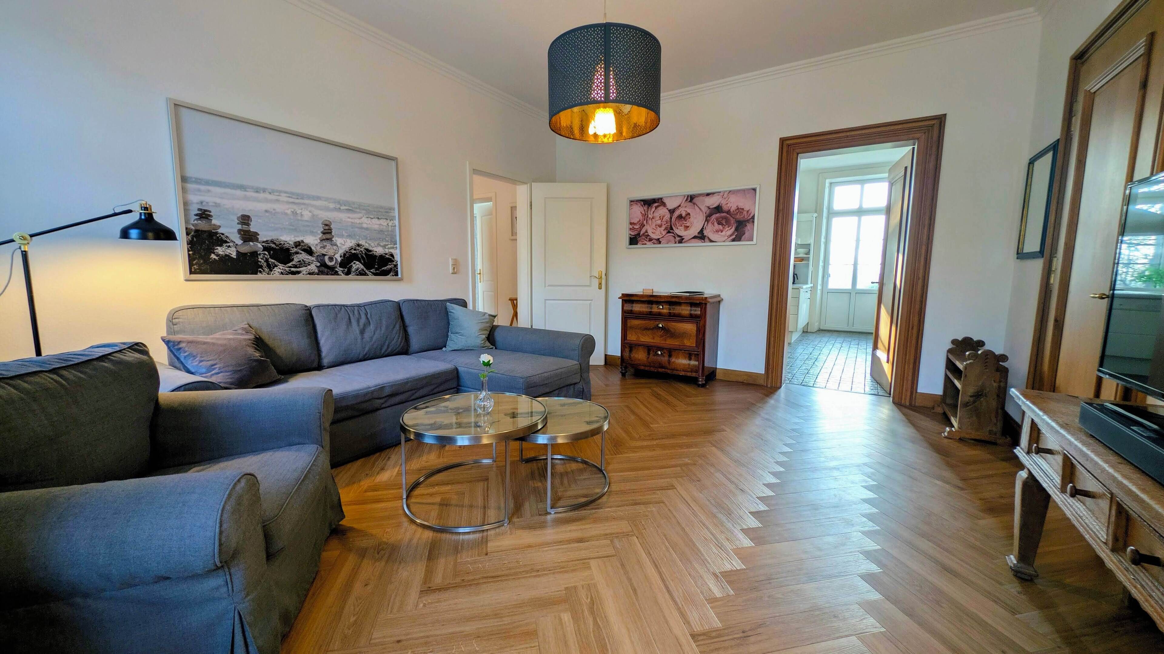 A living room with a grey sofa, a glass coffee table, wooden flooring, a hanging light fixture, and a framed picture on the wall.