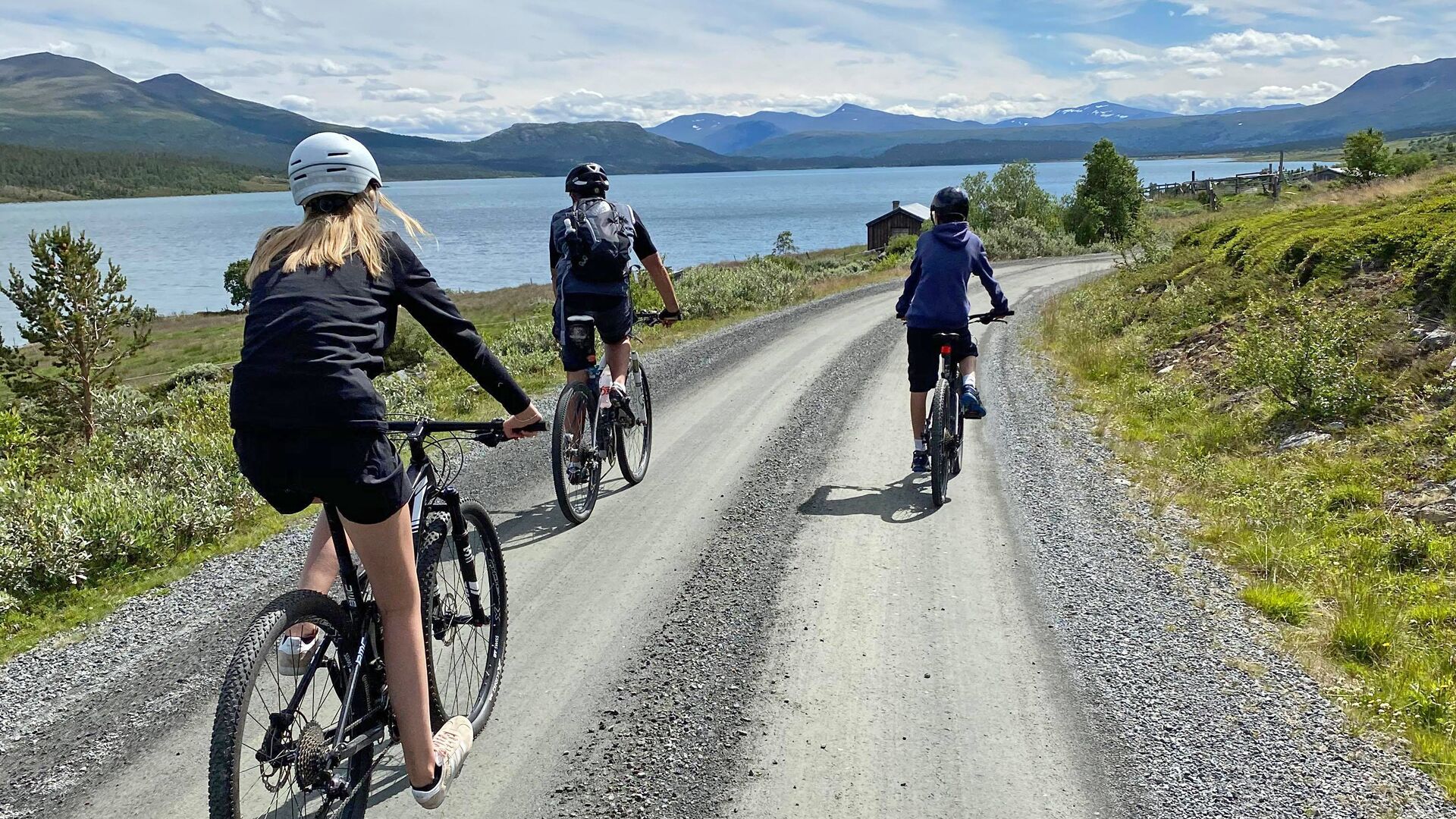 Three cyclists on a gravel road with a lake and mountains in the background.