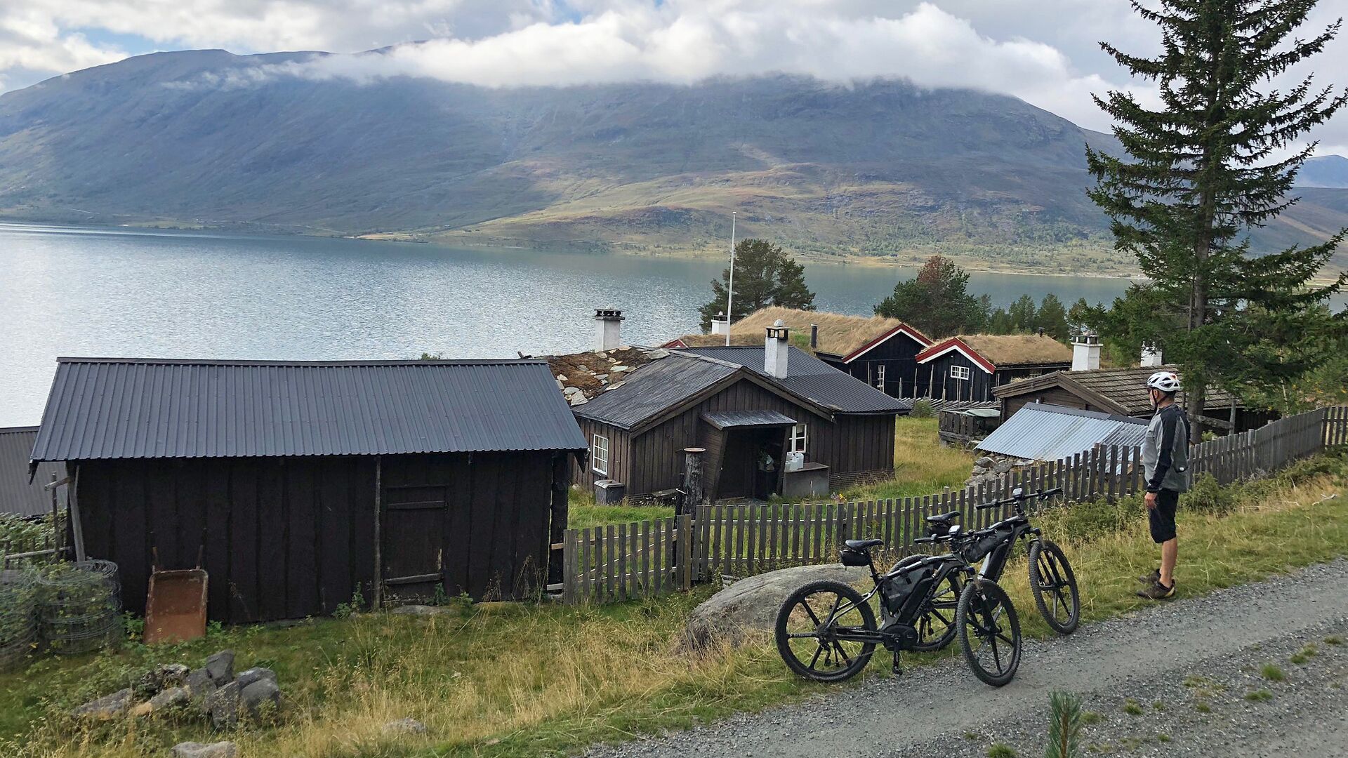 A rural setting with a lake, wooden buildings, and a person with a bicycle.