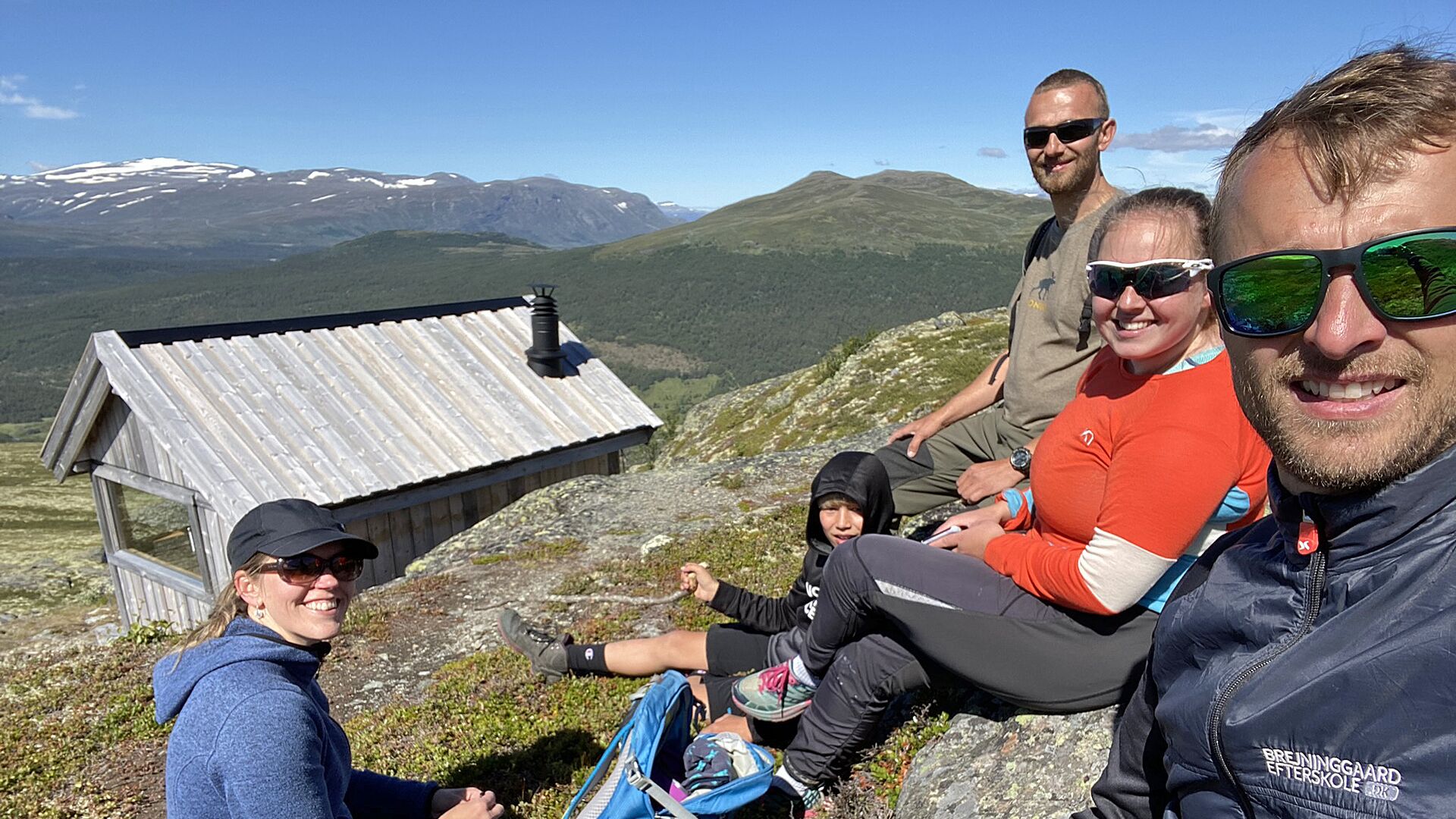 A group of people sitting on a mountain ridge with a small wooden shelter and a view of snow-capped mountains in the background.
