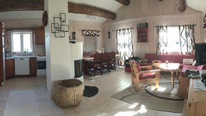 A spacious living area with a dining table, chairs, and a sofa. A kitchen with wooden cabinets and a window is visible in the background.