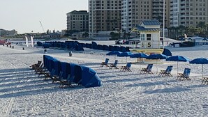 A beach with rows of blue beach chairs and umbrellas, a lifeguard tower, and high-rise buildings in the background.