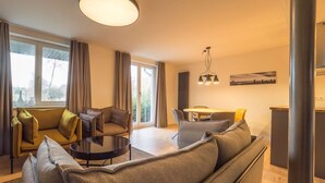 Una sala moderna con un sofá, una mesa de centro y una zona de comedor con sillas. Hay un ventanal con cortinas, una lámpara de techo e imagen colgada en la pared.