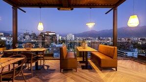 A rooftop terrace with wooden flooring, tables, chairs, and a sofa. There are hanging pendant lights and a view of a cityscape with mountains in the background. - Hotel Nodo - El primer hotel de negocios en Chile (Santiago)