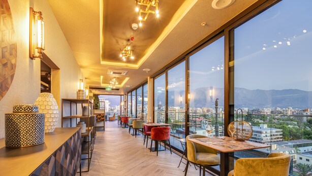 A modern restaurant with a long counter, multiple tables, and chairs. Large windows offer a city view. - Hotel Nodo - El primer hotel de negocios en Chile (Santiago)