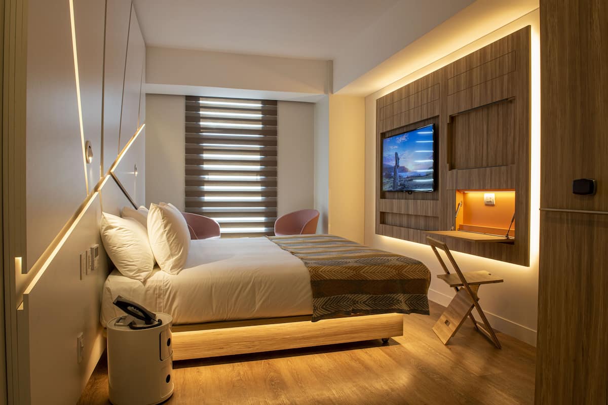 Un dormitorio moderno con cama, televisor montado en la pared, ventana con persianas y una mesita.