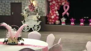 A banquet hall with round tables covered in white tablecloths and chairs with pink sashes. A decorative carriage with pink and white balloons is visible in the background. - The Kingstanding Inn (Birmingham)