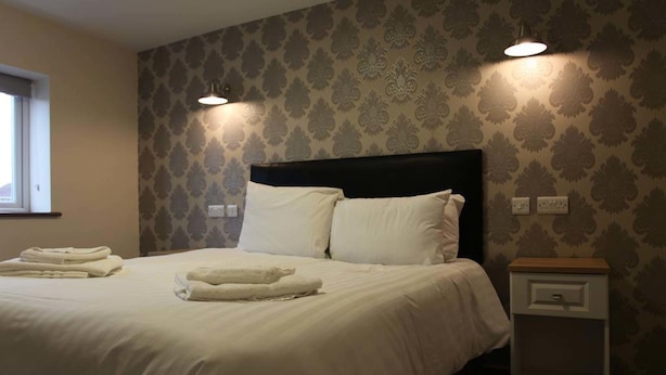 A neatly made bed with white linens and pillows, a bedside table with a drawer, and patterned wallpaper. - The Kingstanding Inn (Birmingham)