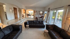 A spacious living area with black leather sofas, a dining table, and a view of the garden through large windows.