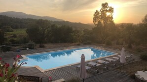 A rectangular swimming pool with a wooden deck, surrounded by lounge chairs and umbrellas, set against a backdrop of trees and hills during sunset.