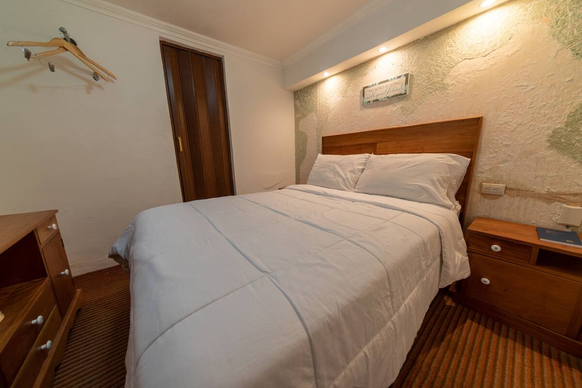 A neatly made bed in a room with a wooden headboard, a wooden nightstand with drawers, a wooden dresser, and a wooden hanger on the wall.