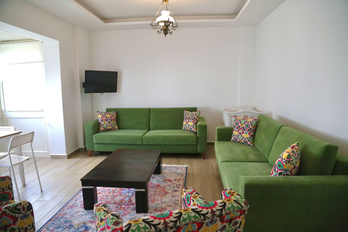 A living room with a green sofa, a coffee table, colorful patterned cushions, a flat-screen TV mounted on the wall, and a chandelier.